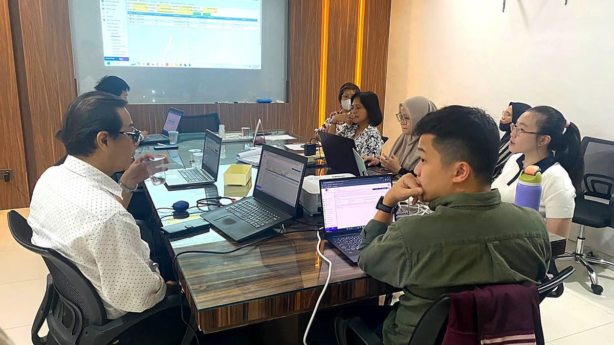 Delapan peserta duduk mengelilingi meja rapat dengan laptop, menghadap layar proyektor yang menampilkan aplikasi vCloudProperty+ (tampilan layar diburamkan).